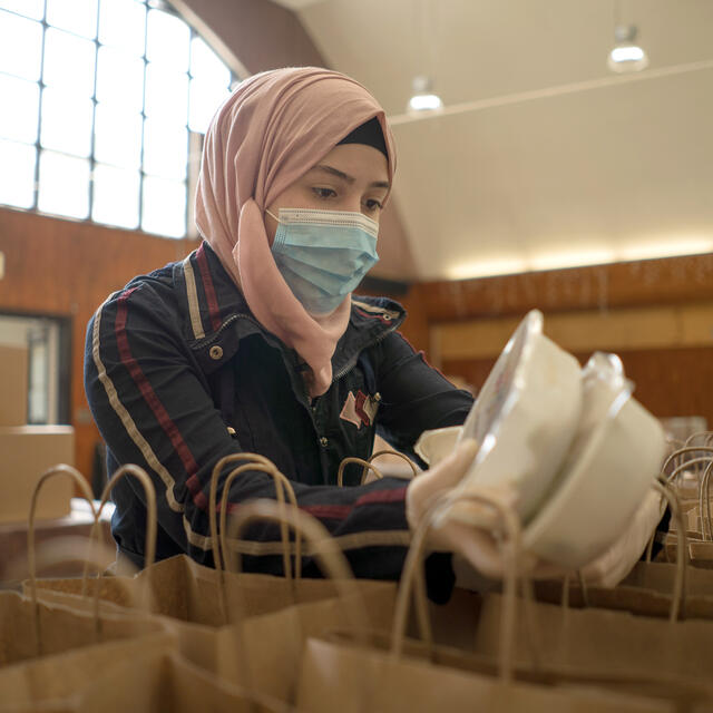 Rania Abou, a 19-year-old refugee from Damascus, Syria, distributes meals to people experiencing food insecurity in Elizabeth, New Jersey. Rania Abou, wearing a mask, stands behind a table filled with paper bags and adds pre-packaged meals to them.