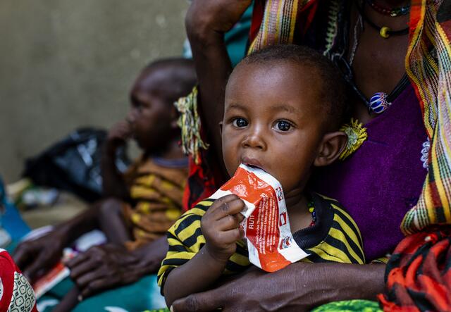 a child licking on the packaging of some emergency food while sitting on it's mothers legs