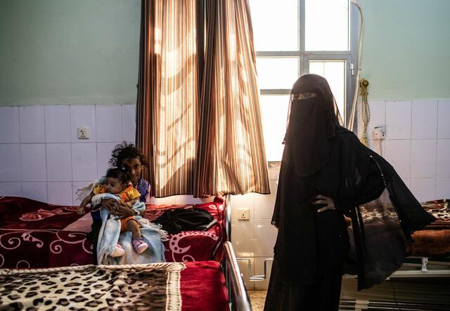a little girl and a baby sit on a bed in a clinic - a fully veiled woman stands beside them