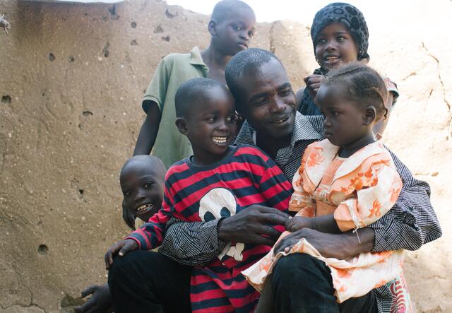 Some children sitting on Simon. Most are smiling except the youngest.