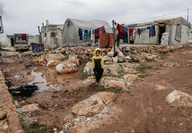 A young boy stands in a rocky and muddy road between baracks and tents