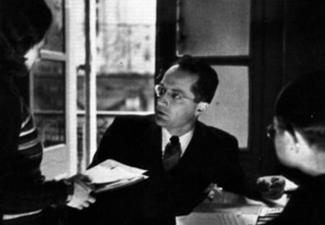 Ein schwarz-weiß Bild. Ein Mann mit einer Brille sitzt an einem Schreibtisch