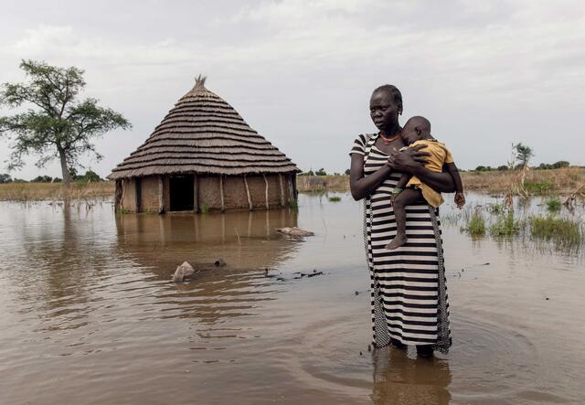 A woman and her child are standing in the water. Behind them is a hut.