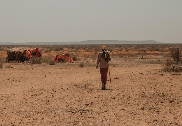 A man carrying a staff walks through a parched landscape in Ethiopia amid makeshift tents with mountains in the distance under a hazy sky.