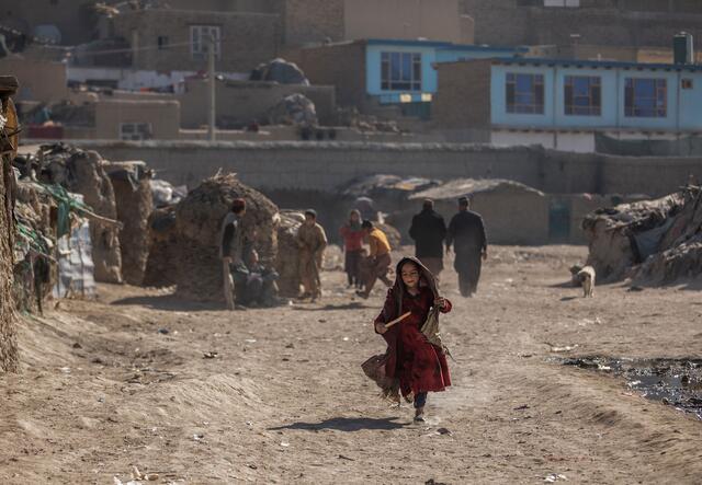 A girl in a red dress runs through a camp for displaced people in Kabul, Afghanista. There are people an makeshift homes in the background. 