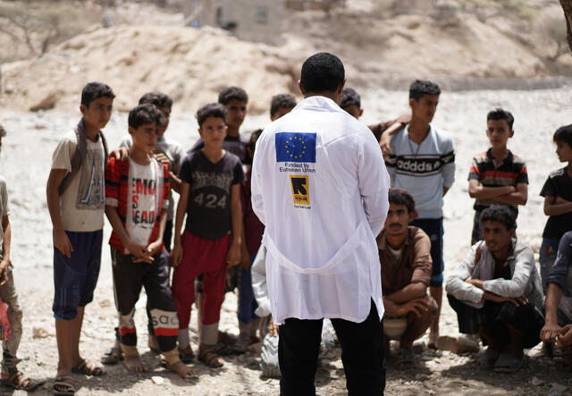 Doctor in a white coat with EU flag on the back stands in front of a line of patients