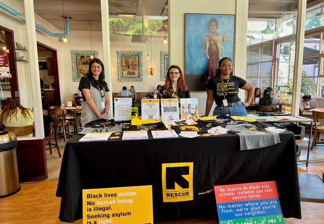 Three young women stand behind a table of IRC in Atlanta t-shirts, water bottles, and other marketing materials.