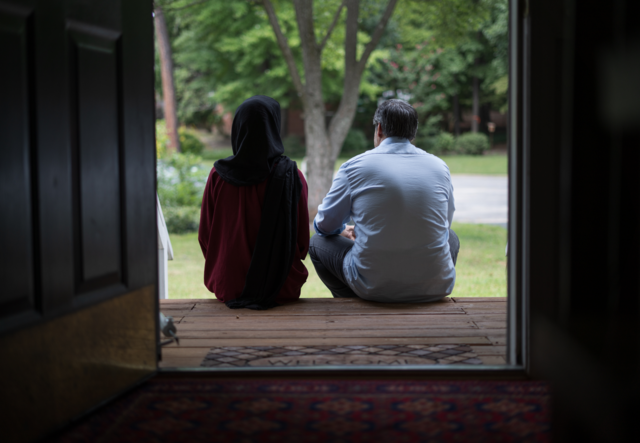 A man and wife sit with their back to the viewer and look at their front lawn.