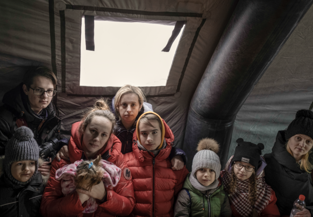 Eight Ukrainian refugees gather in a tent at the Medyka border crossing point in Poland. They all wear warm winter jackets.