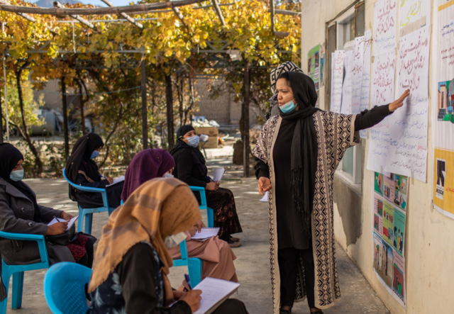 A woman instructs other women who are sitting and taking notes