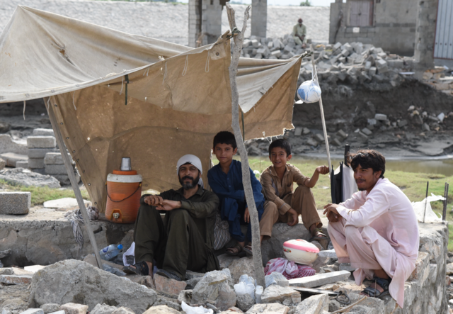 A man, a teenager and two boys sit under a frail makeshift tent. In the background is a pile of rubble which used to be their home.