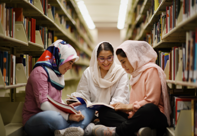 아리파, 자흐라, 하디사는 도서관 한가운데에 원을 그려 함께 앉아있습니다. 함께 책을 읽으며 웃고 있습니다.