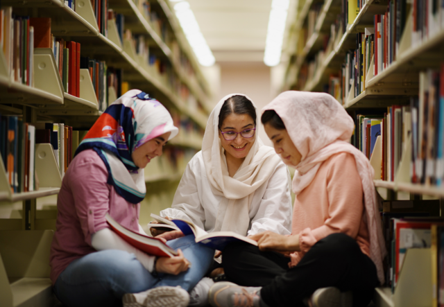 Arifa, Zahra and Hadisa sit together in a circle in the middle of a library. They smile while reading together,