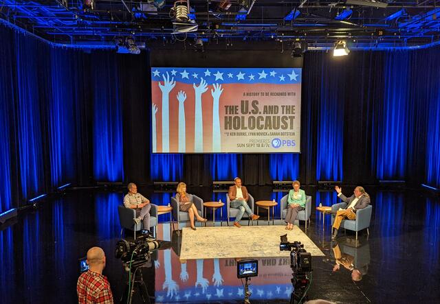 A panel of refugee and immigration experts sit in a row in a large auditorium, with Executive Director of the IRC in Atlanta, Justin Howell, on the far left. Around them are stage lights, film cameras, and a projection of 'The US and the Holocaust' promotion card.