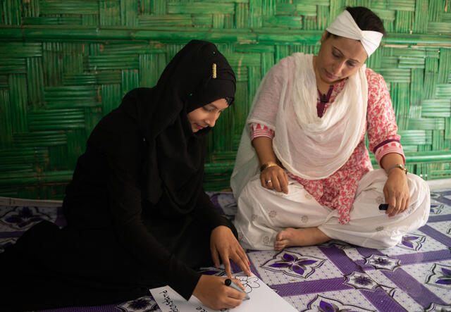 Two women sit on the floor. One of them draws a flower on a piece of paper while the other watches.