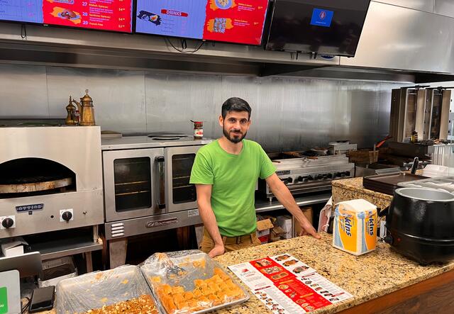 Khaled Abo Alhawa standing in the kitchen of Kababji Grill. Behind him is are several TV screens displaying menu items, a stove, ovens, and a fryer while he leans on a granite countertop.