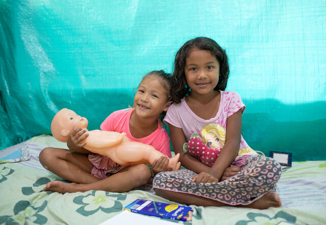 Two girls sit together smiling and holding dolls.