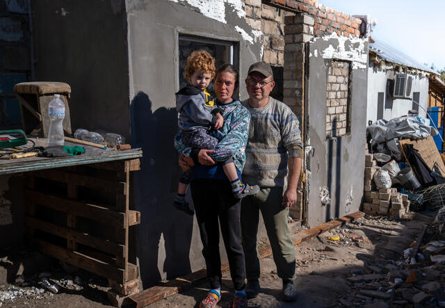 Marina stands next to her husband while holding their son. In the background is their home that was destroyed by the war.