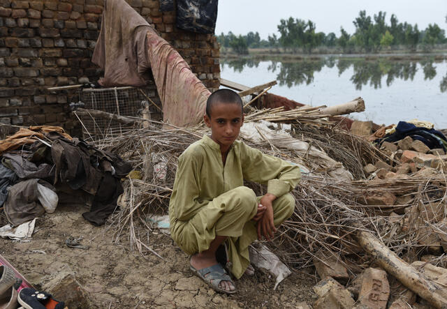Faisal, a young boy, sits outside in the ruins of his family home. In the background, a field is submerged by floodwater.