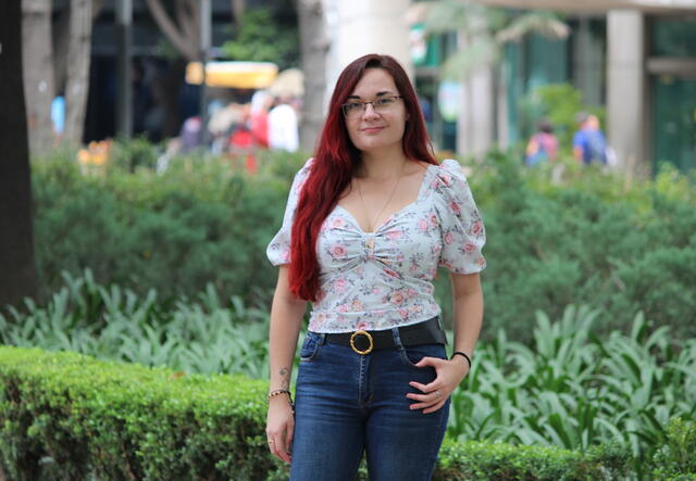 A woman stands in front of greenery with a hand on her pocket.