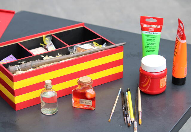 Una caja roja y amarilla con una varita y otros suministros se muestra sobre una mesa junto a recipientes para líquidos y cepillos.