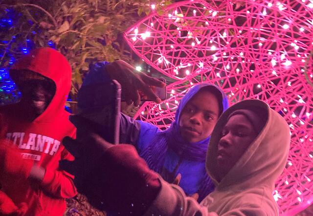 A group of 3 male students holding a phone to take a selfie in front of pink lights.