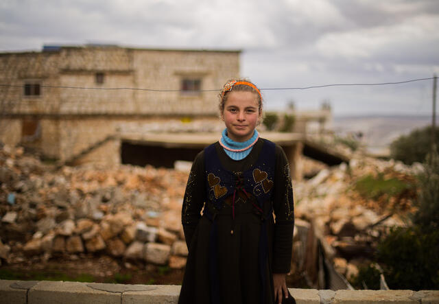 Girl stands in front of family home that has been devastated by an airstrike.