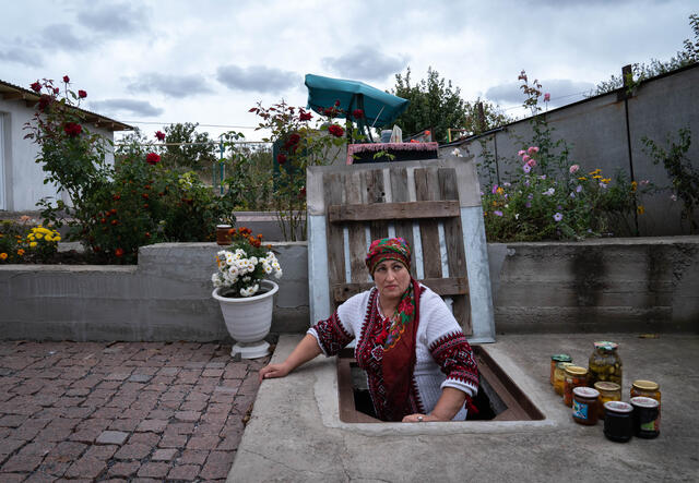 Olga stands on a ladder, half-way standing in her underground cellar. She has taken out a variety of pickled fruits and vegetables which sit in jars on the ground next to her.