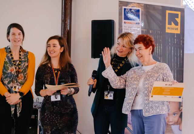 Four women standing next to each other, one holding a certificate and waving