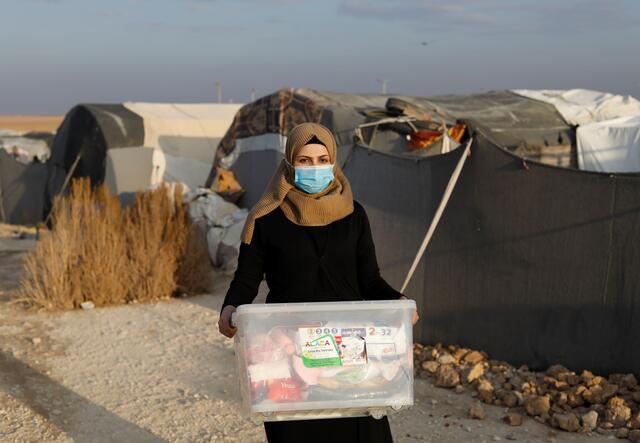 A woman standing in front of tents and holding a plastic box filled with items