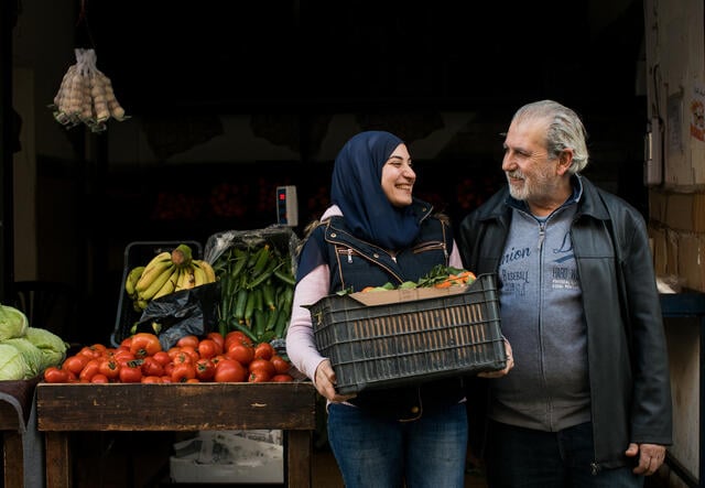 Ala'a and her father stand outside the entrance to her shop, Ala'a holding a box of fruit and veg..