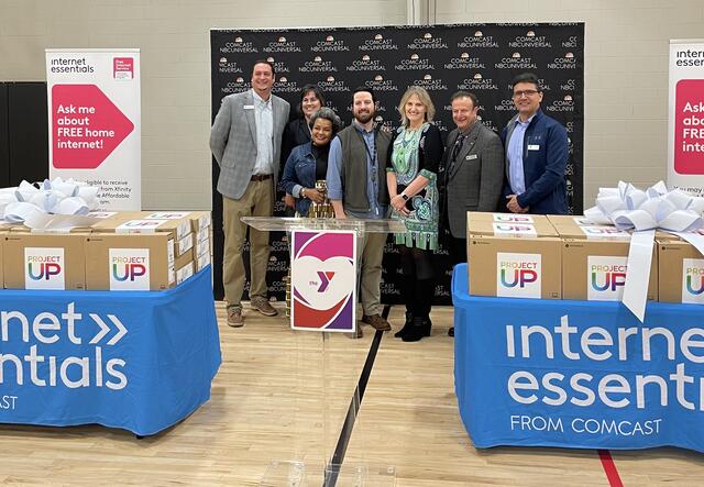 3 women and 4 men stand smiling behind tables with donated laptops