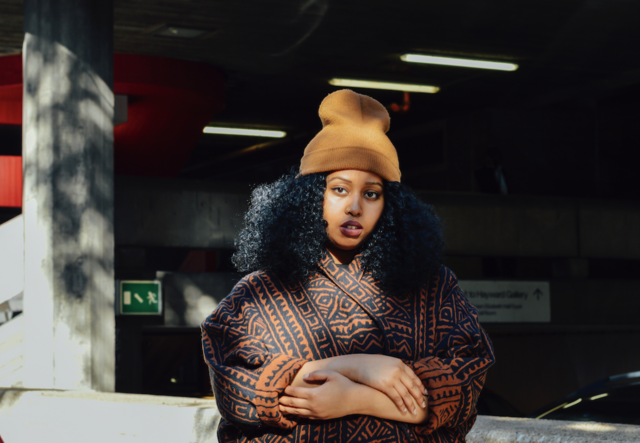 Warsan Shire stands with arms crossed, wearing a yellow hat