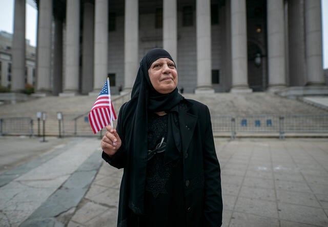 Maha stands outside a Federal office building waving a US flag and smiling.