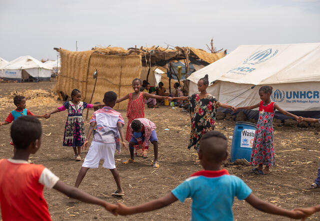 Children play together at Tunaydbah camp, Sudan. Most of the children hold hands and form a circle around two children who complete an activity in the middle.