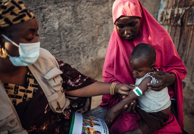 An IRC nutrition officer teaches a mother how to screen her child for signs of malnutrition using MUAC tape in Nigeria. The malnutrition wraps the tape around a boys arm while the mother looks on.