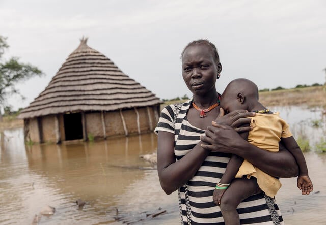 Abuk hält ihr Kind Nyirou vor ihrem überfluteten Haus. Das Wasser hat das Gebiet rund um das Haus überflutet.