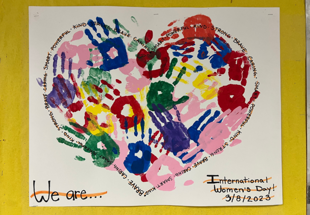 A collage of painted handprints in the shape of a heart outlined with messages of female empowerment for International Women's Day.