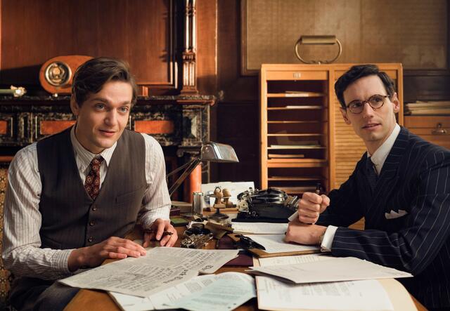 Still from Netflix series "Transatlantic" - Lucas Englander as Albert Hirschmann and Cory Michael Smith asVarian Fry sit at a table together, facing the camera.