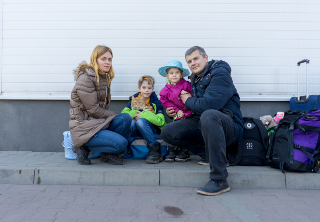 Parents, two children and a cat pose for a photo. Besides them are suitcases packed with their belongings.