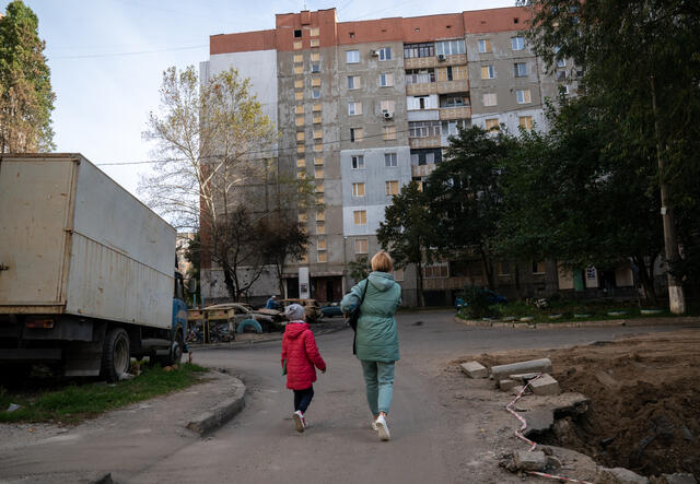 A mother and her child walk home past a crater caused by a missile strike.