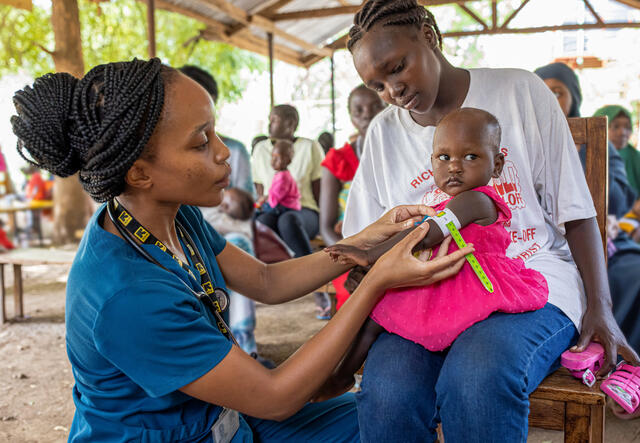 케냐의 야외 의료 센터에서 국제구조위원회 직원이 엄마의 무릎에 앉은 어린 소녀의 영양실조 징후를 검사합니다.