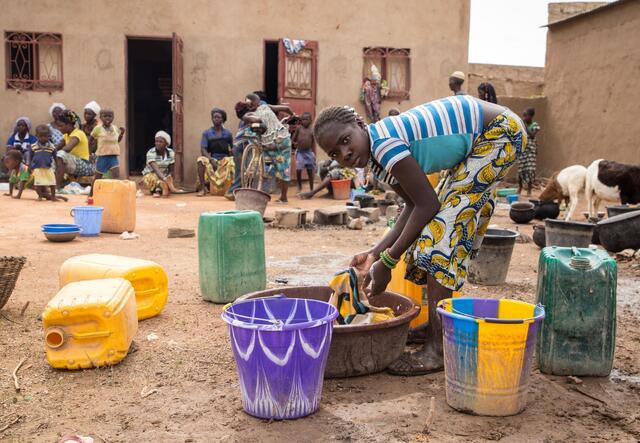 A girl washes clothes in a plastic bucket outside of a building.