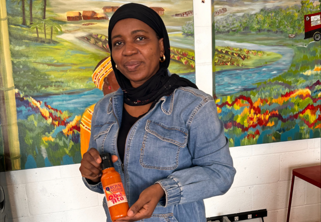 Nimota Dairo standing and holding a bottle of her very own Royal Nigerian Foods sauce. She is wearing a black head scarf and a blue demin jacket.