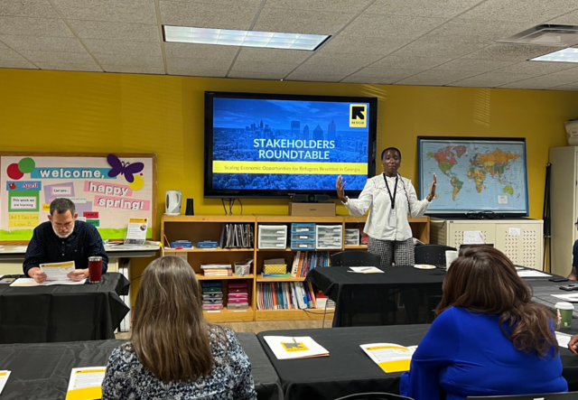 Senior Development Manager, Heloise Ahoure, leading the presentation for the stakeholders roundtable. To her right is Executive Director of the IRC in Atlanta, Justin Howell, and in the foreground are two female attendees sitting at a table with their backs to the camera.