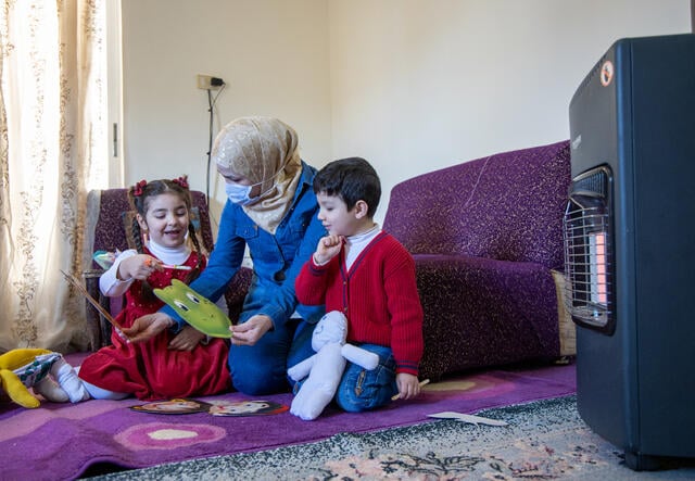 Sulaima and her two children sit on the floor of their house. The children are playing with educational toys from the Ahlan Simsim program.