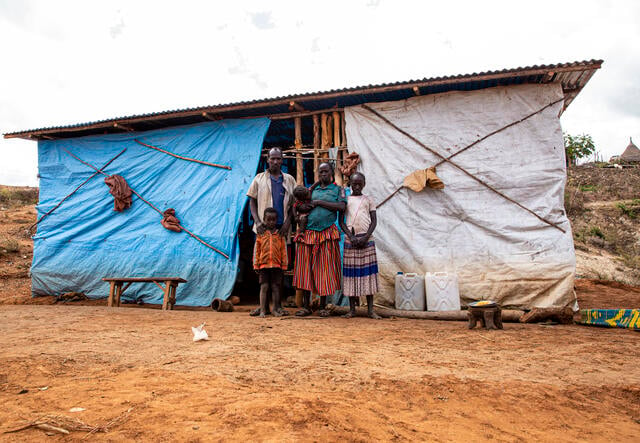 Gedeno, his wife, and three of their children stand outside of their home in Ethiopia, which seems to be constructed of wooden poles and plastic tarps.