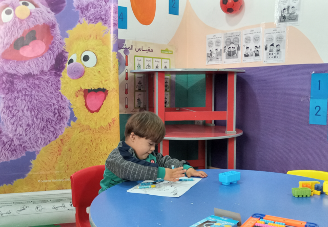 Five year old Saeed draws in a coloring book during an Ahlan Simsim session in Syria. The young boy smiles brightly and points to pictures of his favorite Ahlan Simsim characters.