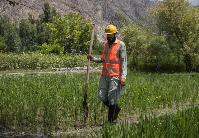 Zhwan Mall walks through a field, carrying a long spade.