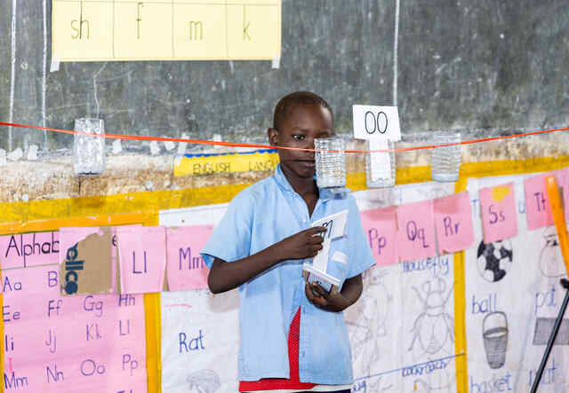A young boy participates in an active learning activity in a Ugandan classroom. The student stands at the front of the room and arranges letters on a line to spell out the word "moon".